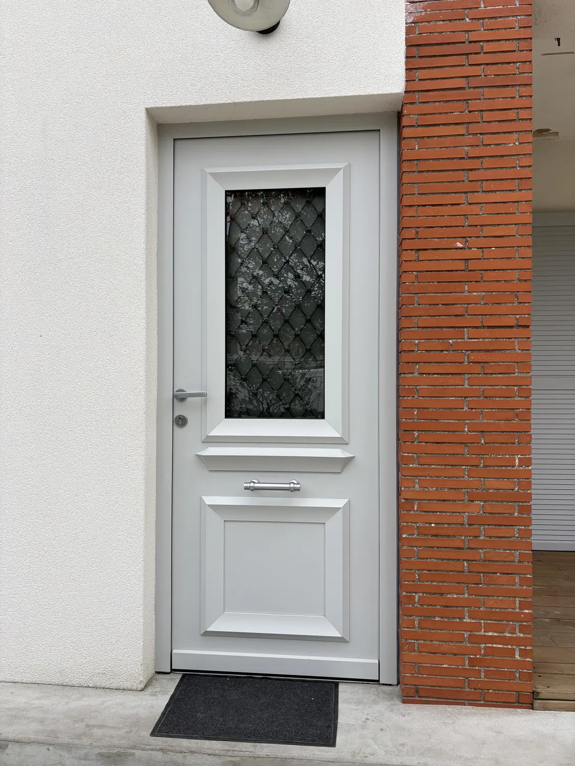 Porte d'entrée semi-vitrée à motif losange entre un enduit clair et un pilier de brique