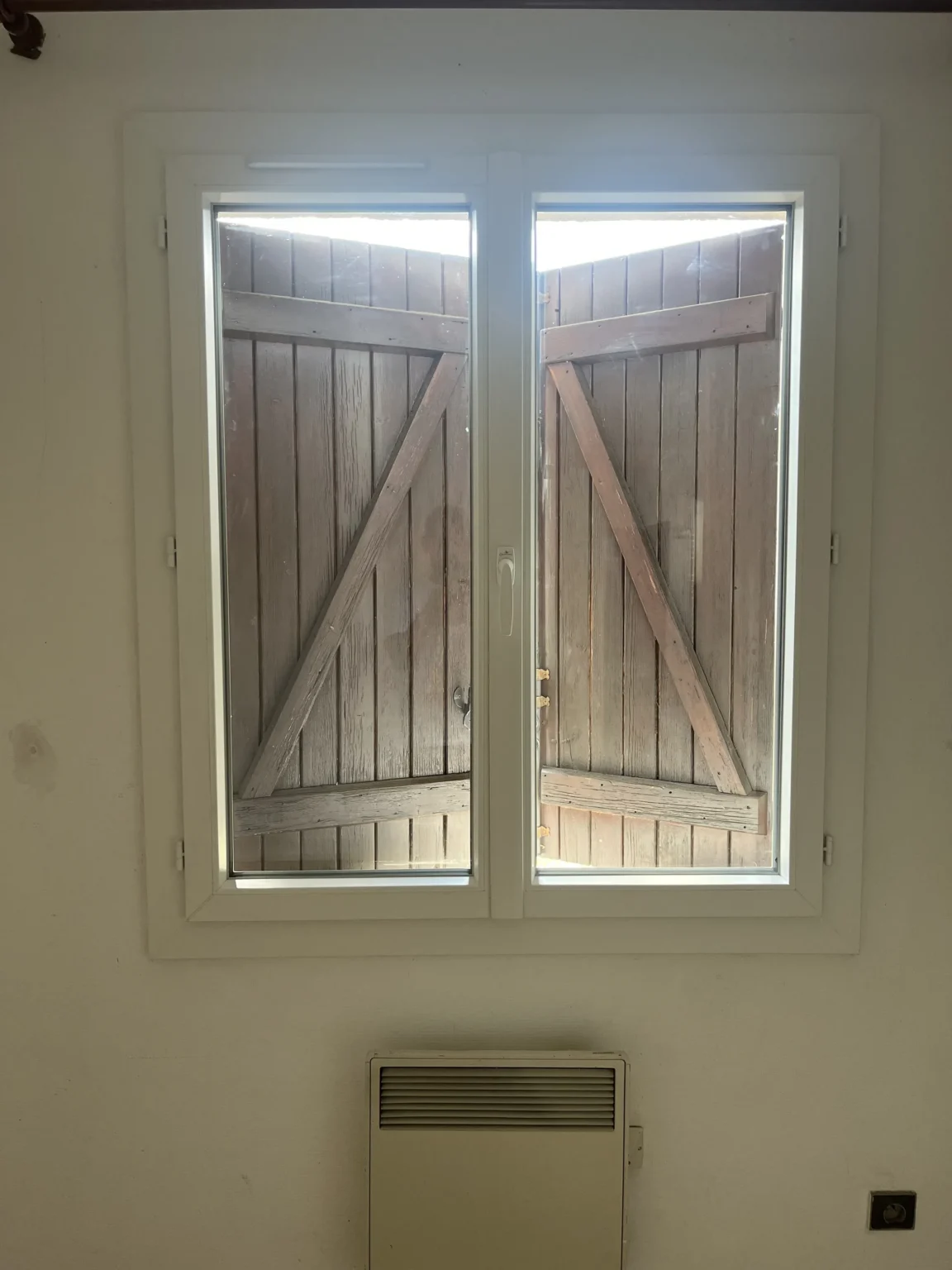 Vue intérieure d’une porte-fenêtre PVC ouvrant sur une terrasse carrelée, volets bois ouverts, Saint-Jean-d’Illac