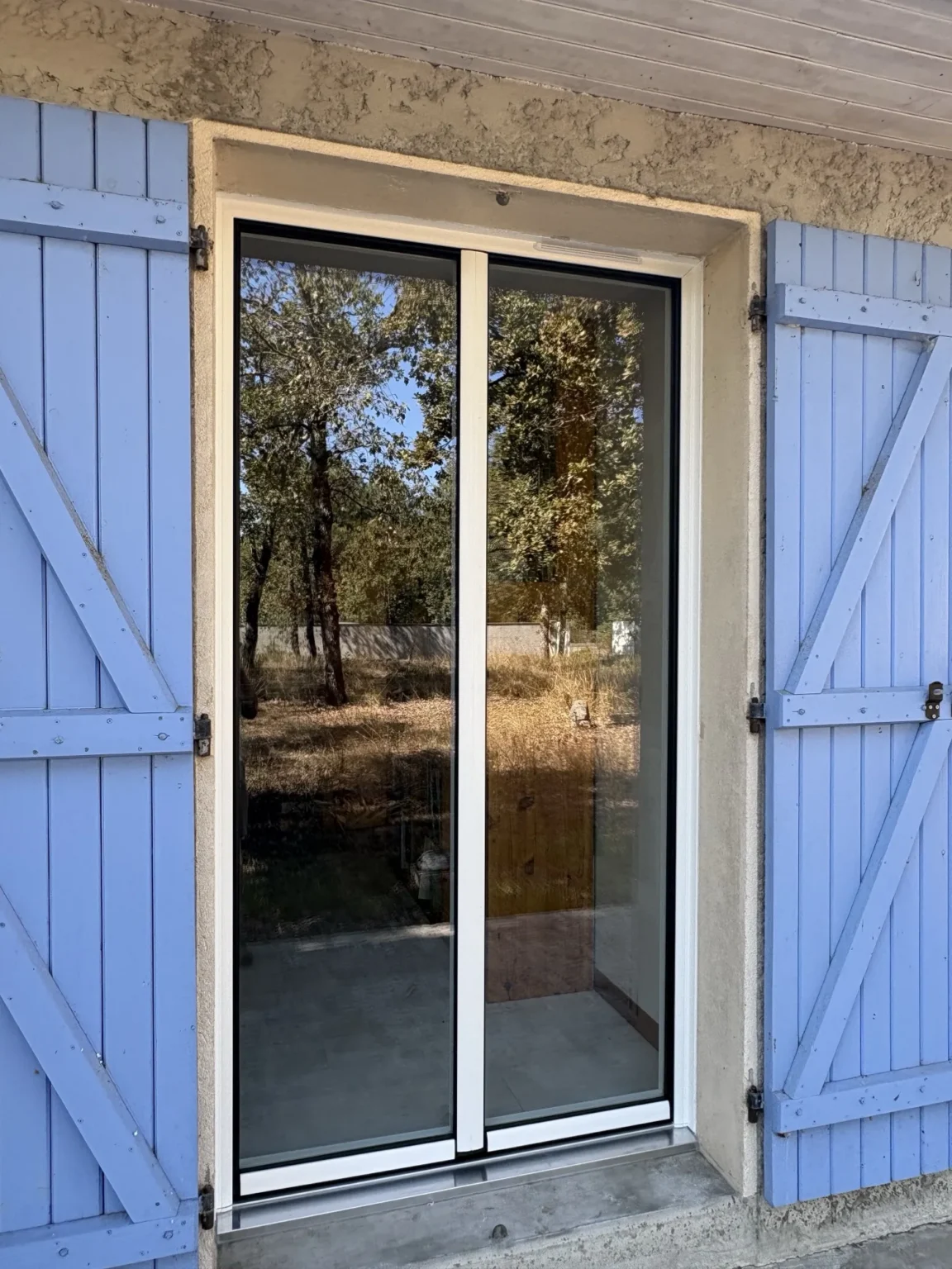 Porte-fenêtre aluminium deux vantaux au profil fin, sous le débord de toit, volets bois bleu lavande conservés